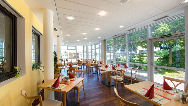Un comedor amplio con múltiples mesas y sillas, ventanas grandes que ofrecen vistas a zonas verdes exteriores y un diseño interior moderno y cuidado.