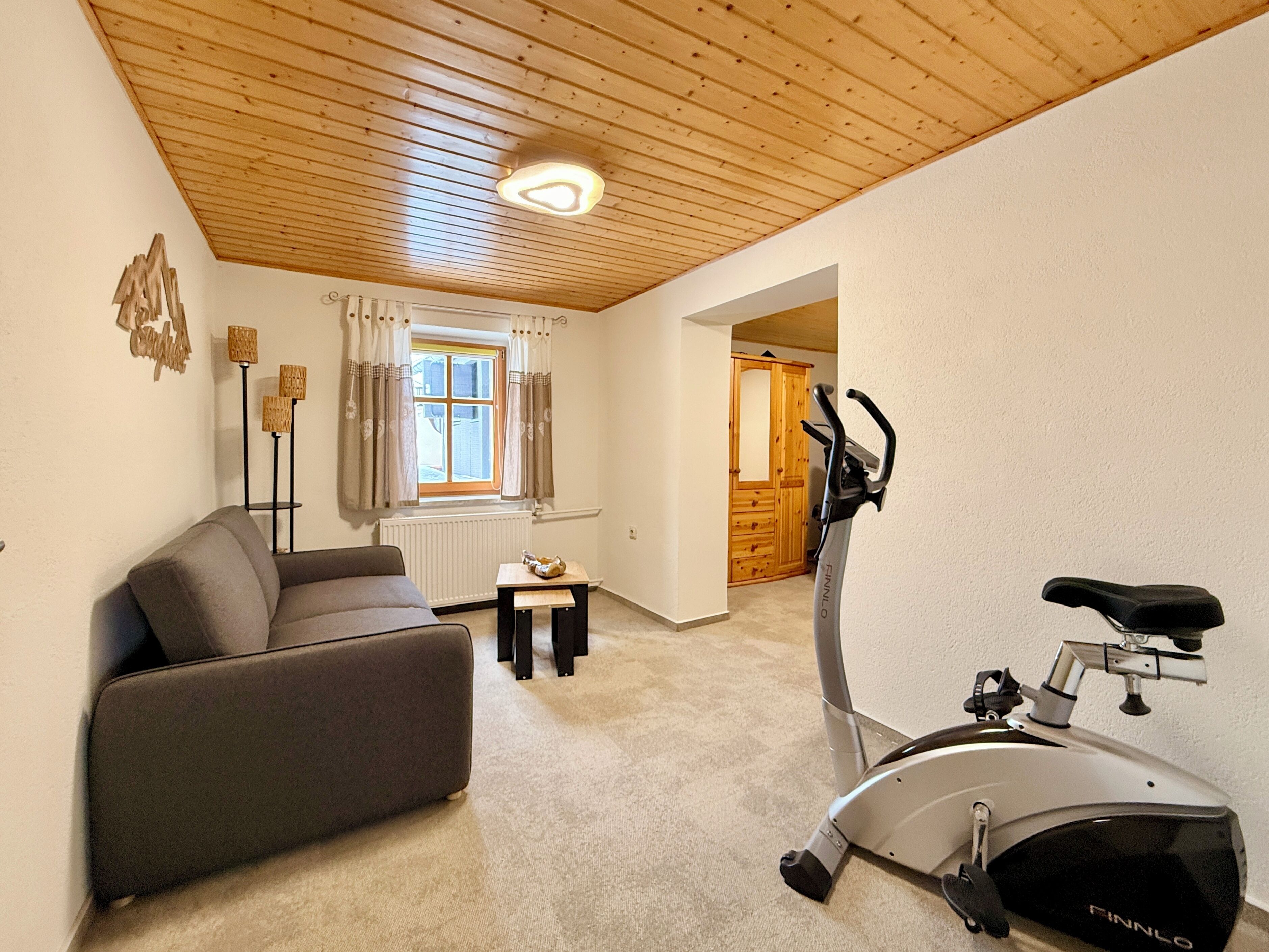 A living room with a sofa, a table with a lamp, a window with curtains, and an exercise bike.