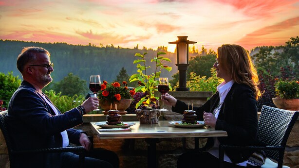 A couple dining at an outdoor table with a view of a sunset and forested hills. - Naturhotel Gasthof Bärenfels (Altenberg)