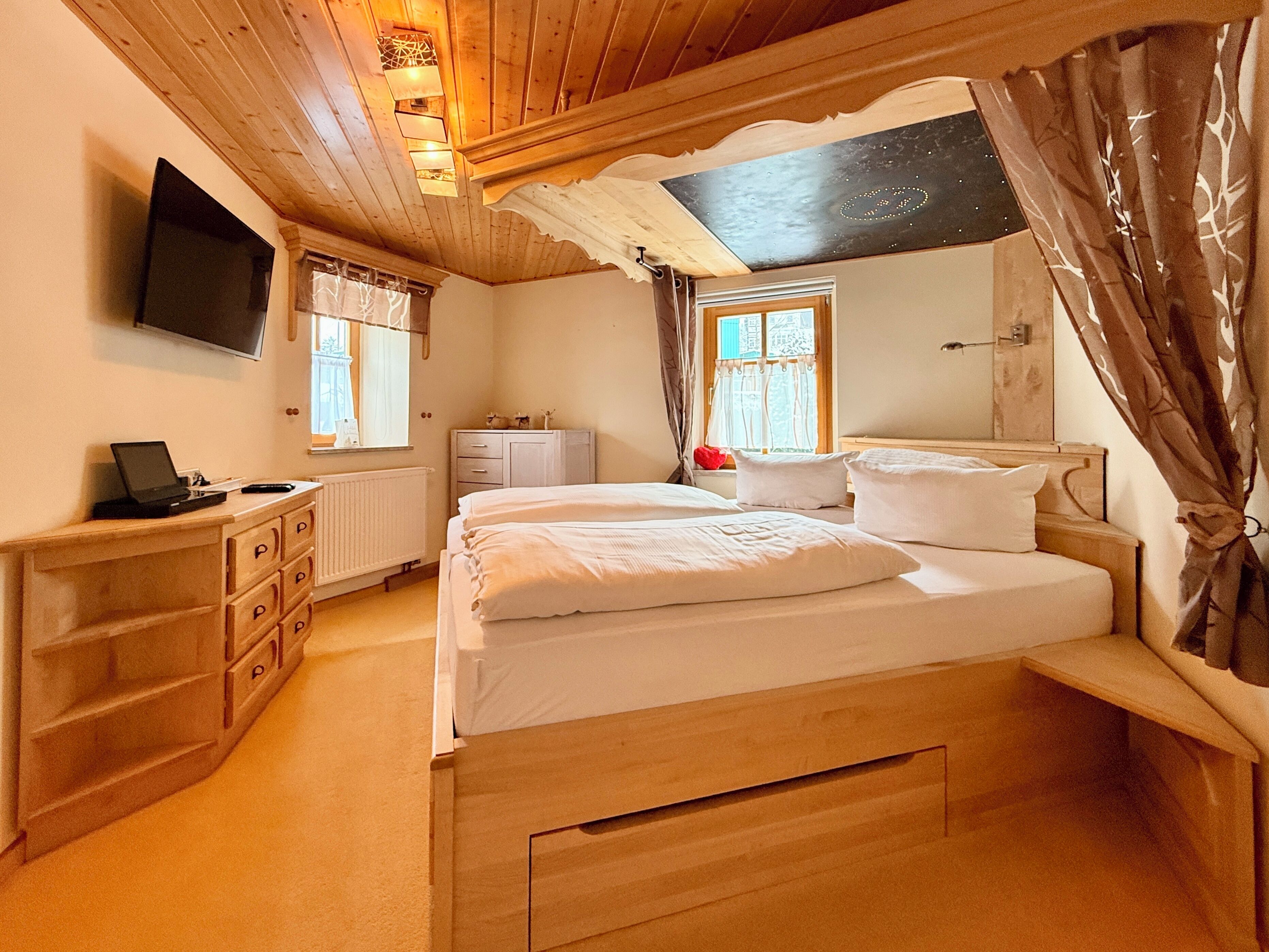 A bedroom with a bed, a desk, a TV, and a window with curtains.
