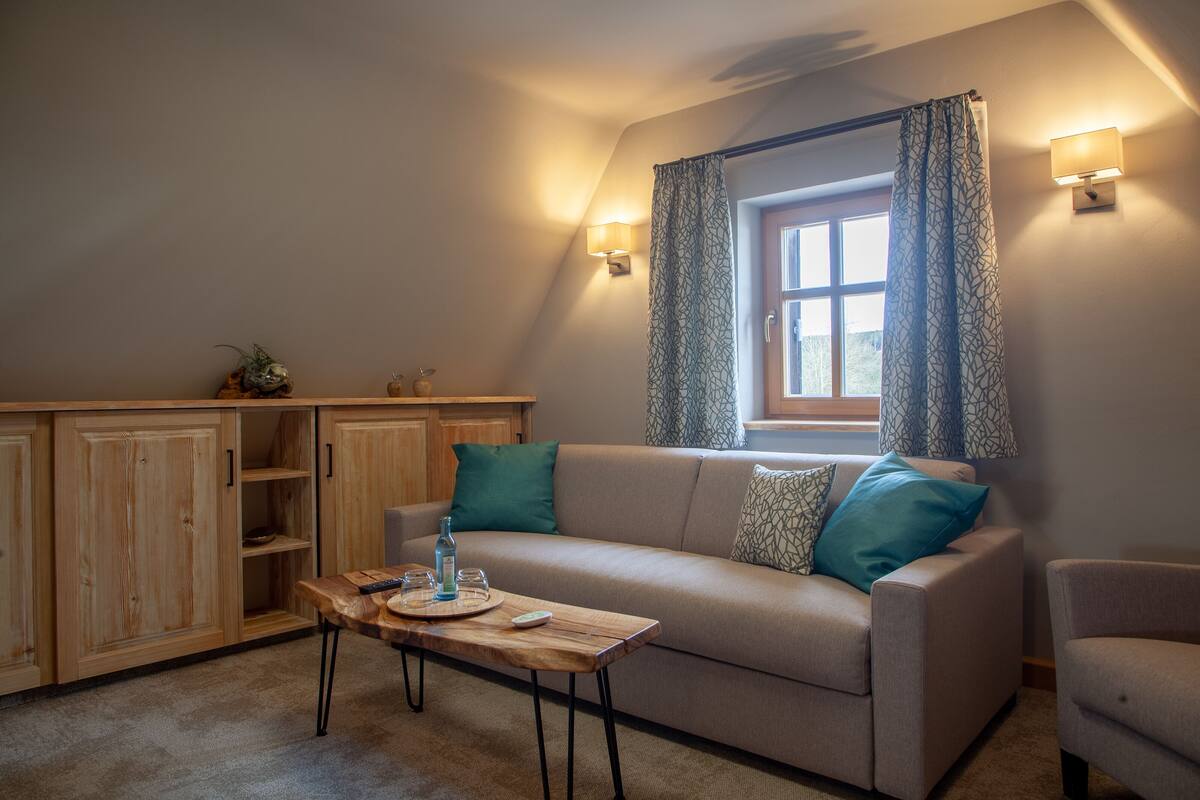 Ein gemütliches Wohnzimmer mit einer Couch, einem Holztisch und einem Fenster mit gemusterten Vorhängen.