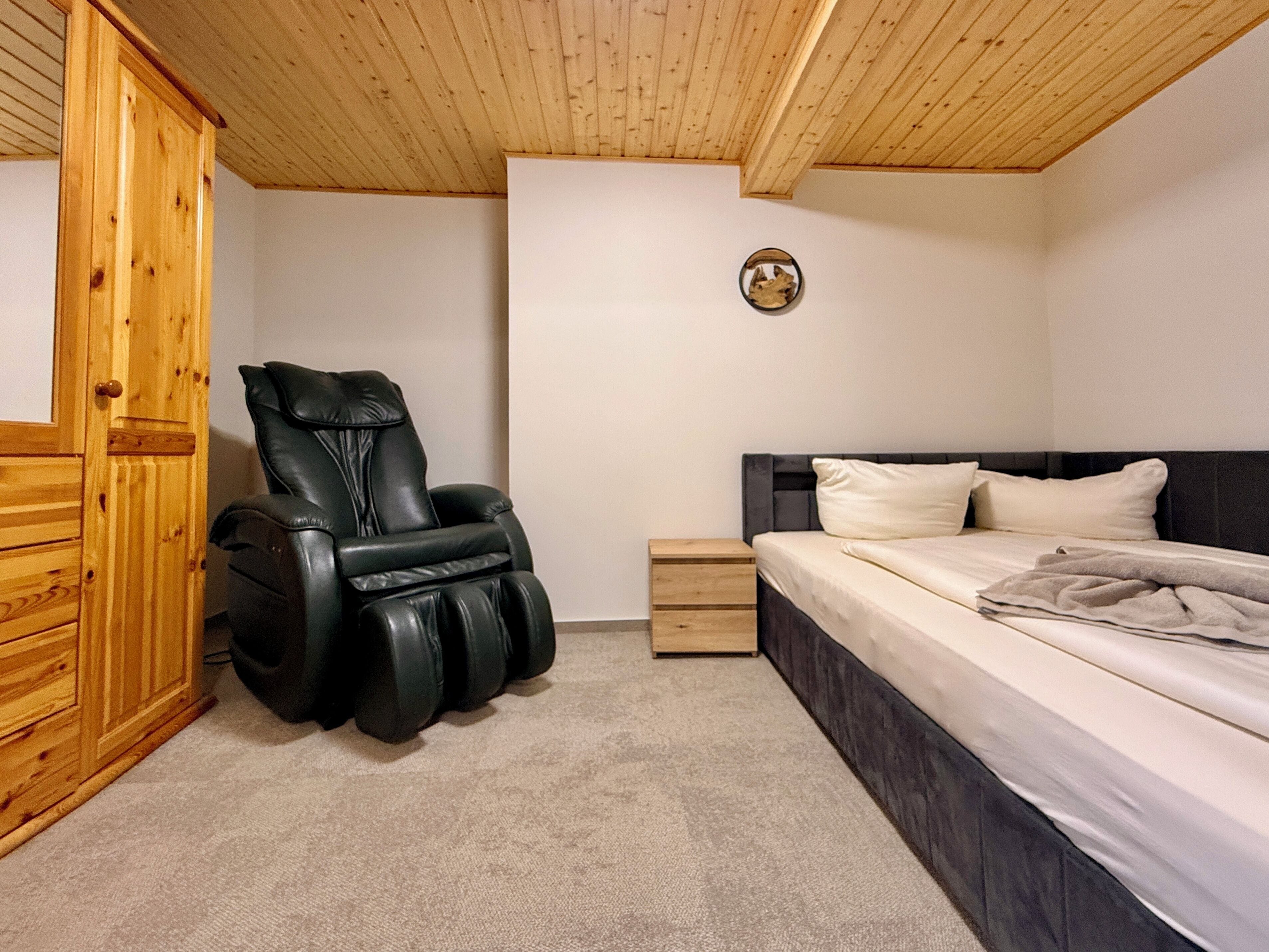 Un dormitorio con sillón de masajes, cama, armario de madera y mesita de noche.