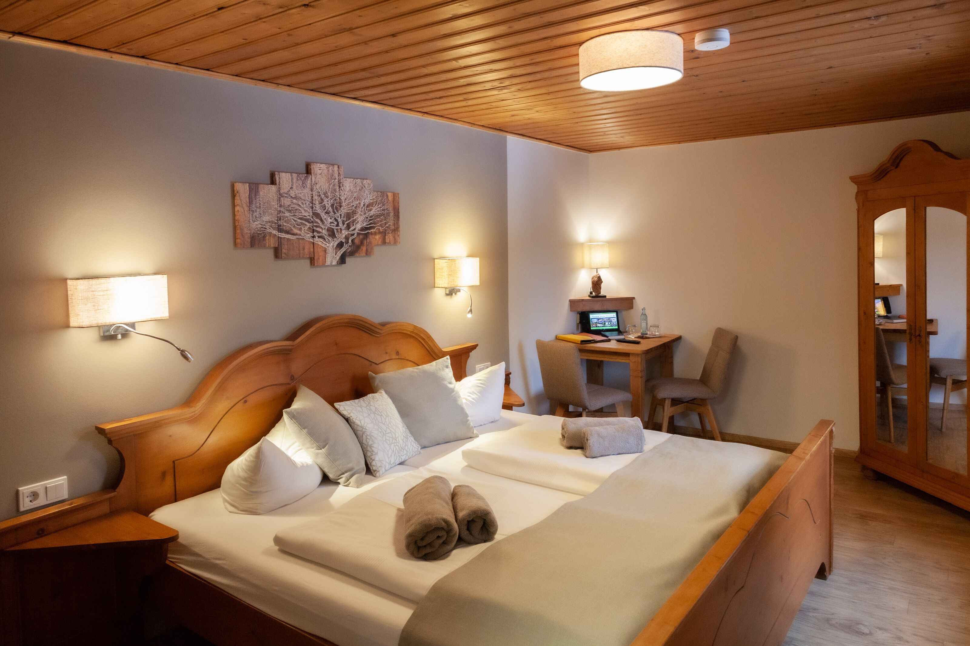 Ein ordentlich bezogenes Bett mit weißen Leinen und aufgerollten Handtüchern, ein Holz-Kopfteil, Nachttischlampen, ein Schreibtisch mit einem Computer und ein Spiegelkorpus.