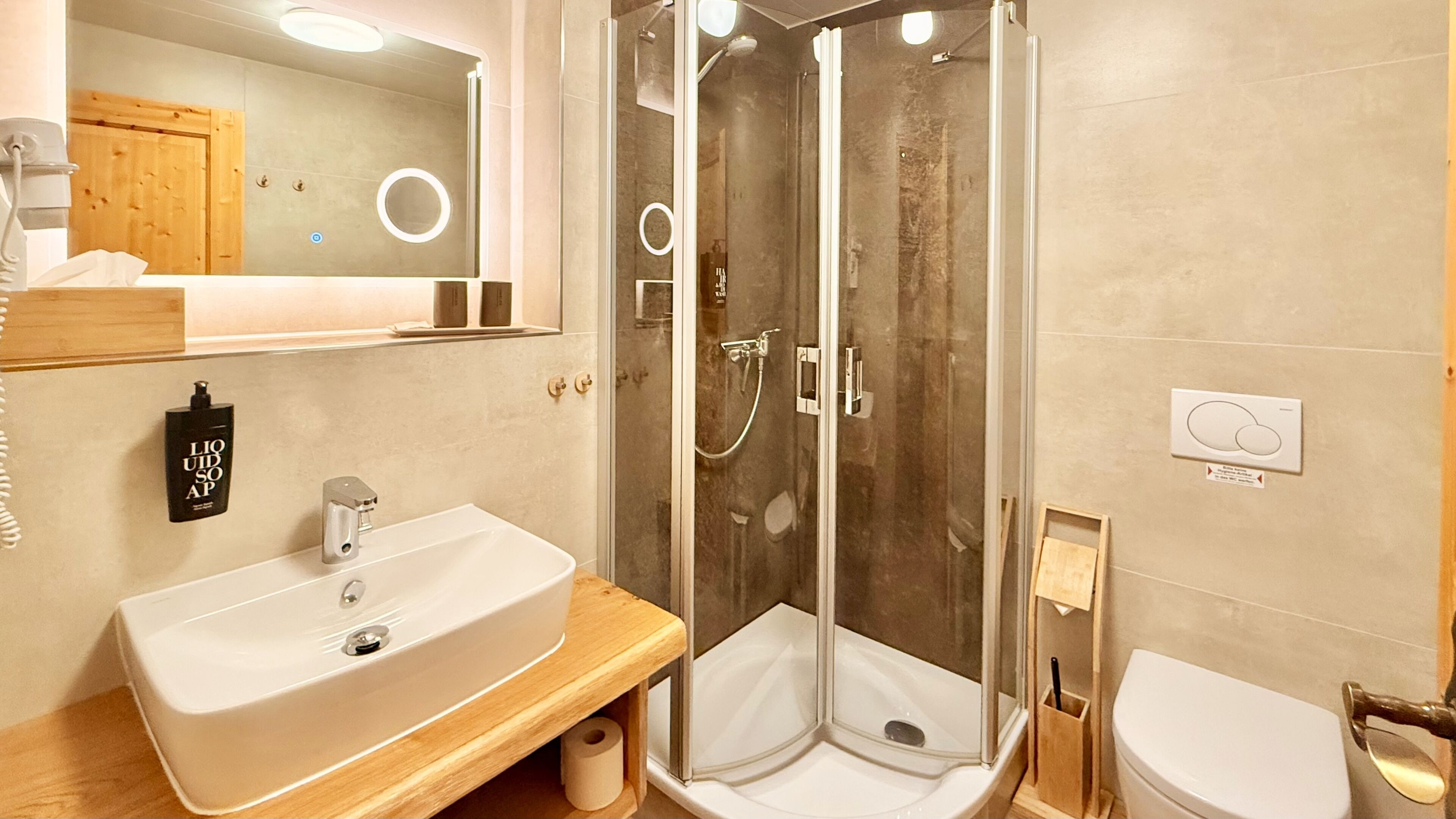 Un bany amb un lavabo blanc, una dutxa amb portes de vidre, un vàter i un mirall amb un armari de fusta a la part superior.