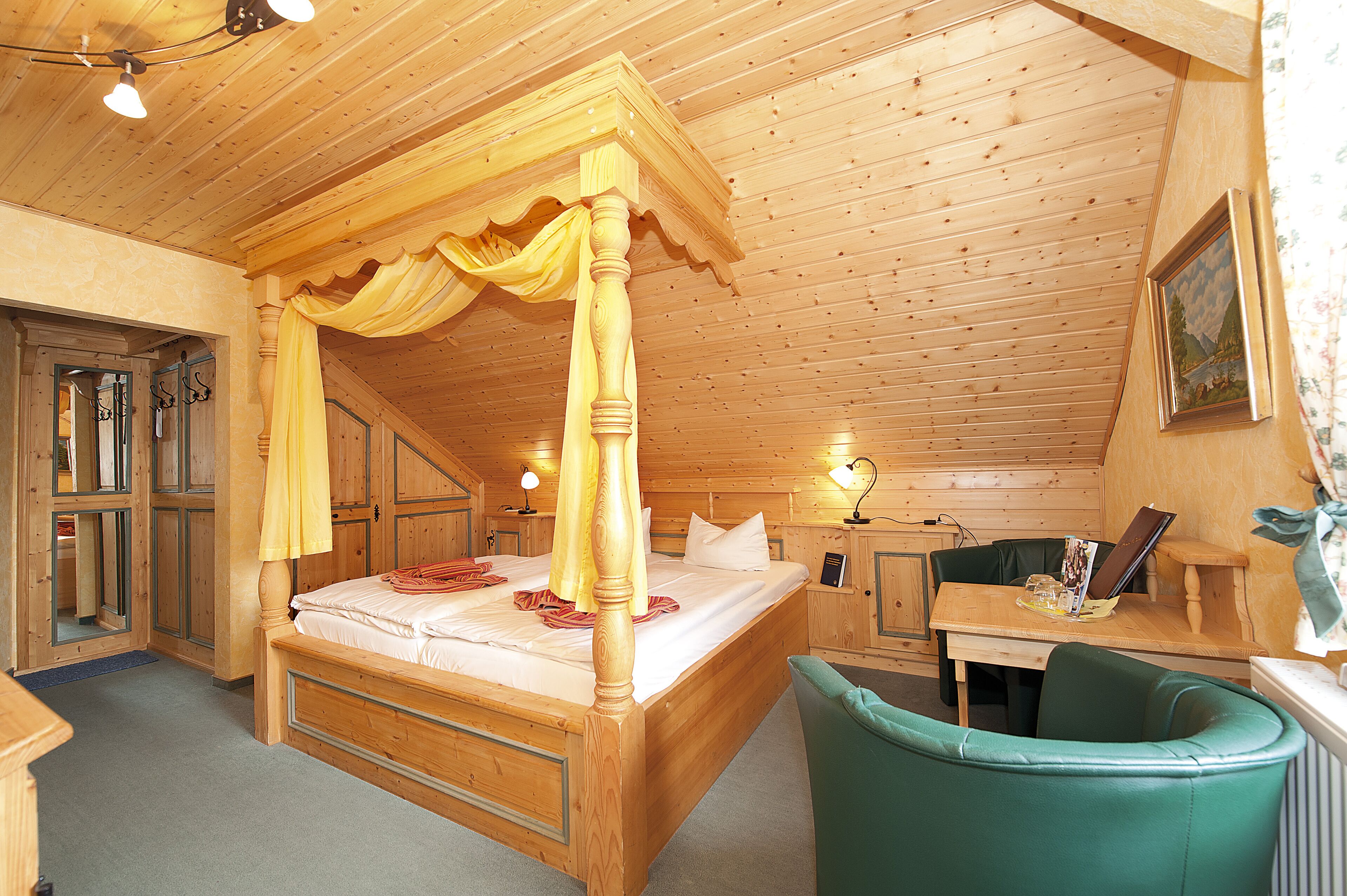 Una cama con dosel de madera con cuatro columnas, un sillón tapizado en verde y un escritorio de madera con una computadora portátil y libros.