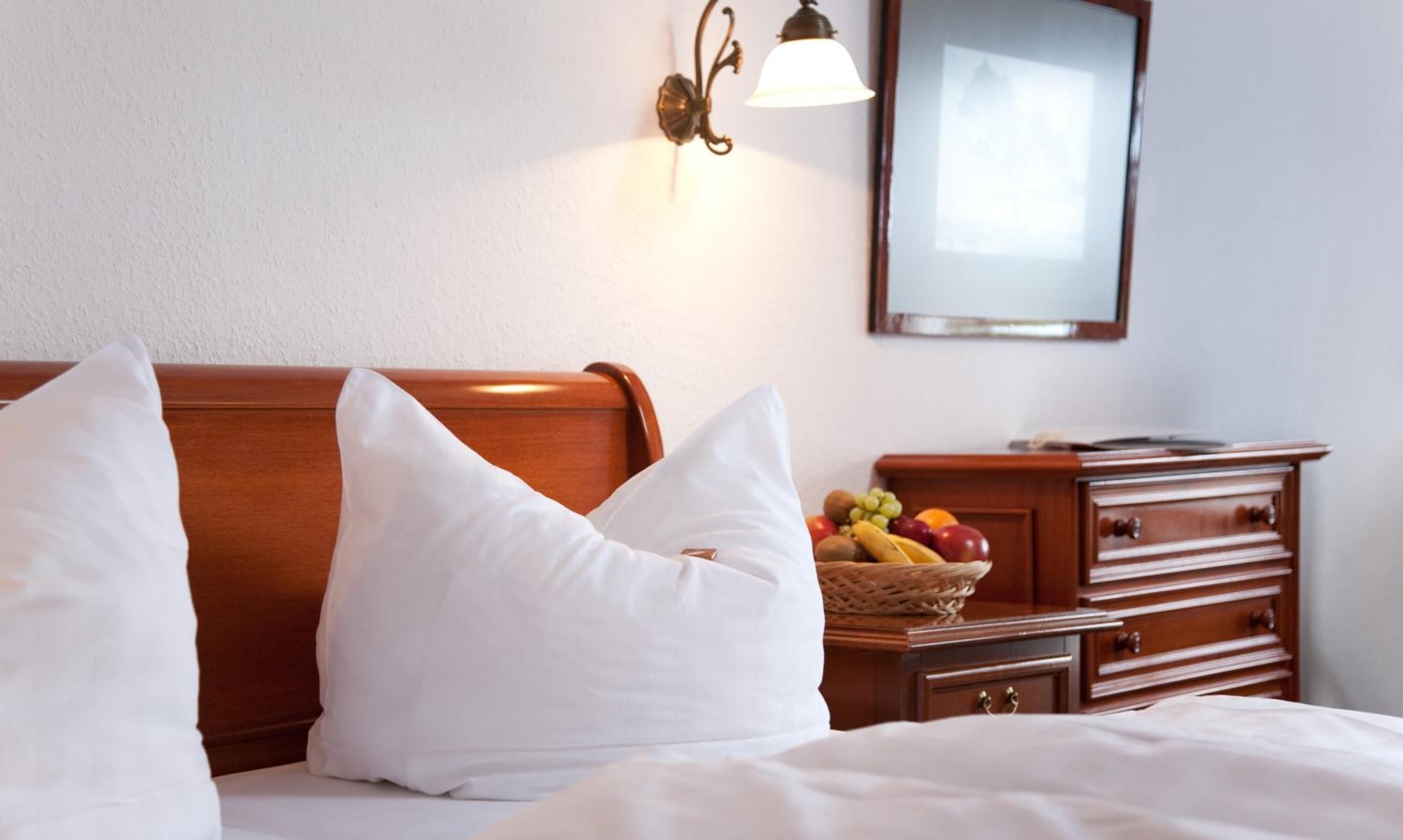 A hotel room with a bed, two white pillows, a wooden nightstand with a fruit basket, and a wall-mounted lamp.