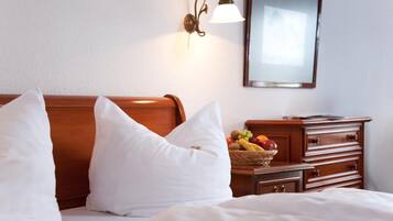 Une chambre d’hôtel avec un lit, deux oreillers blancs, une table de chevet en bois sur laquelle se trouve un panier de fruits, et une lampe fixée au mur.