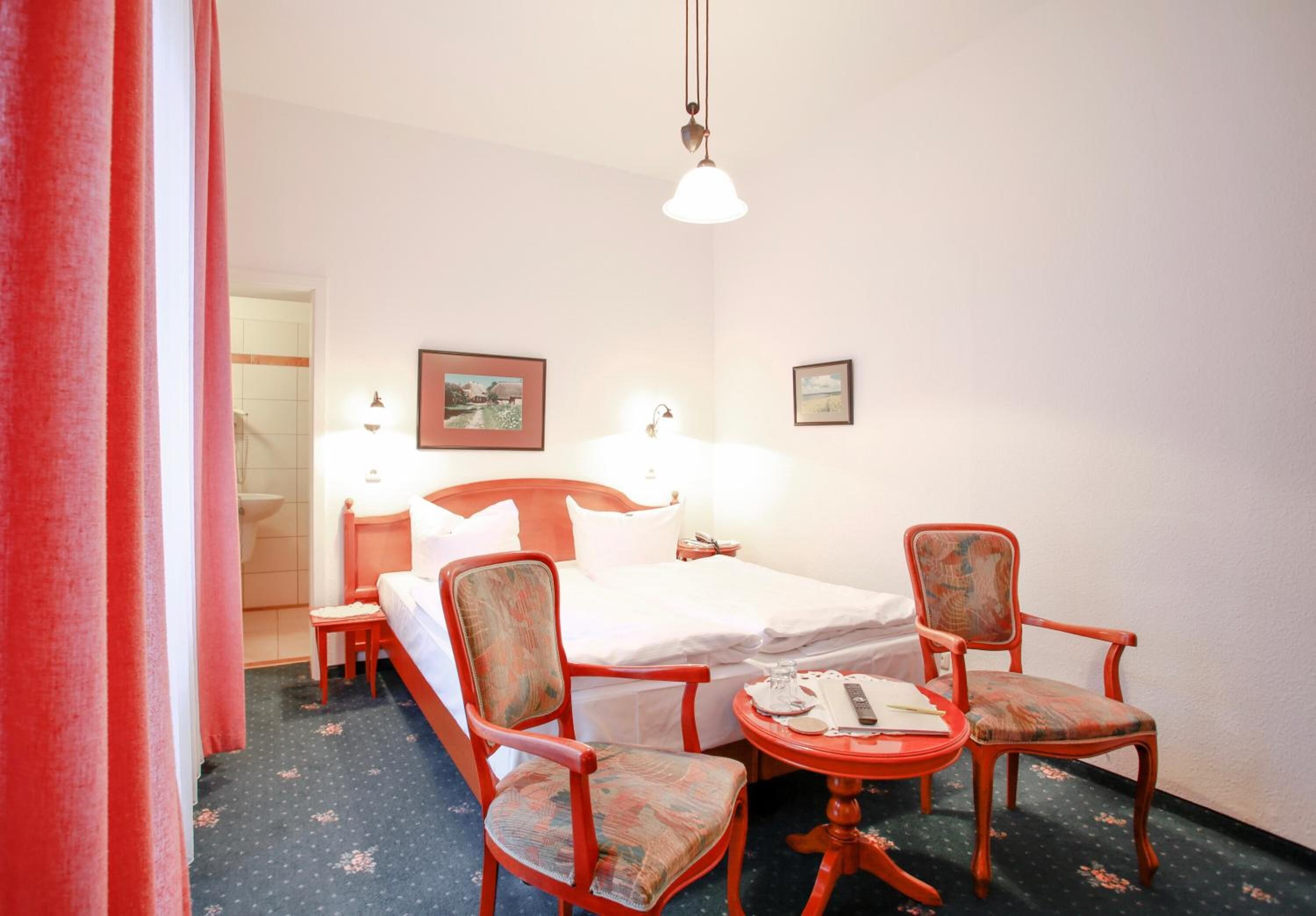 Habitación de hotel con cama, dos sillas, una mesita, lámpara y un cuadro en la pared.