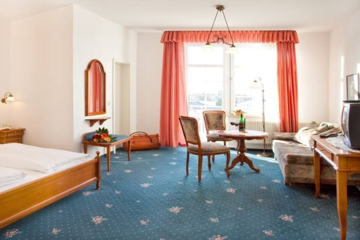 A hotel room with a double bed, a desk with a chair, a small table with a chair, a television, and a window with curtains.