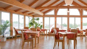 A spacious dining area with wooden tables and chairs, large windows, and potted plants. - Hotel Stranddistel Rügen (Göhren)