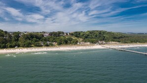 A coastal view with a beach, a pier, and a forested hill. - Hotel Stranddistel Rügen (Göhren)