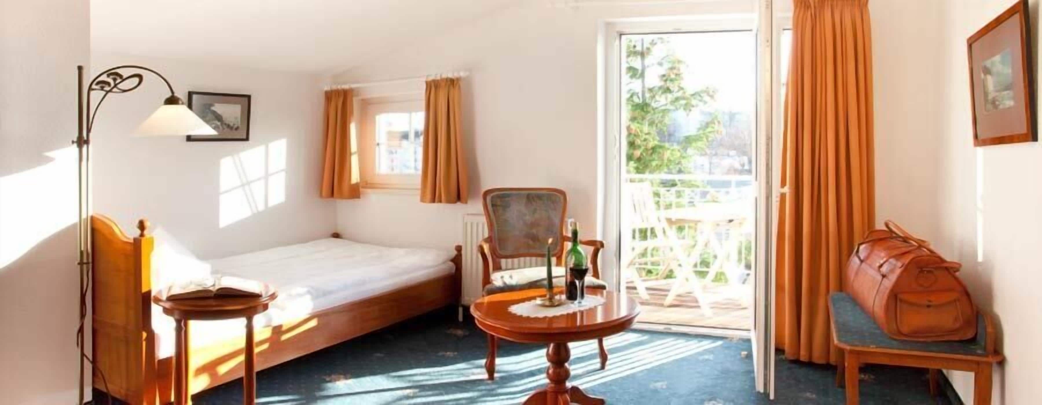 A compact hotel room with a single bed, a small table with a candle, a chair, and a view of the outdoors through a sliding door.