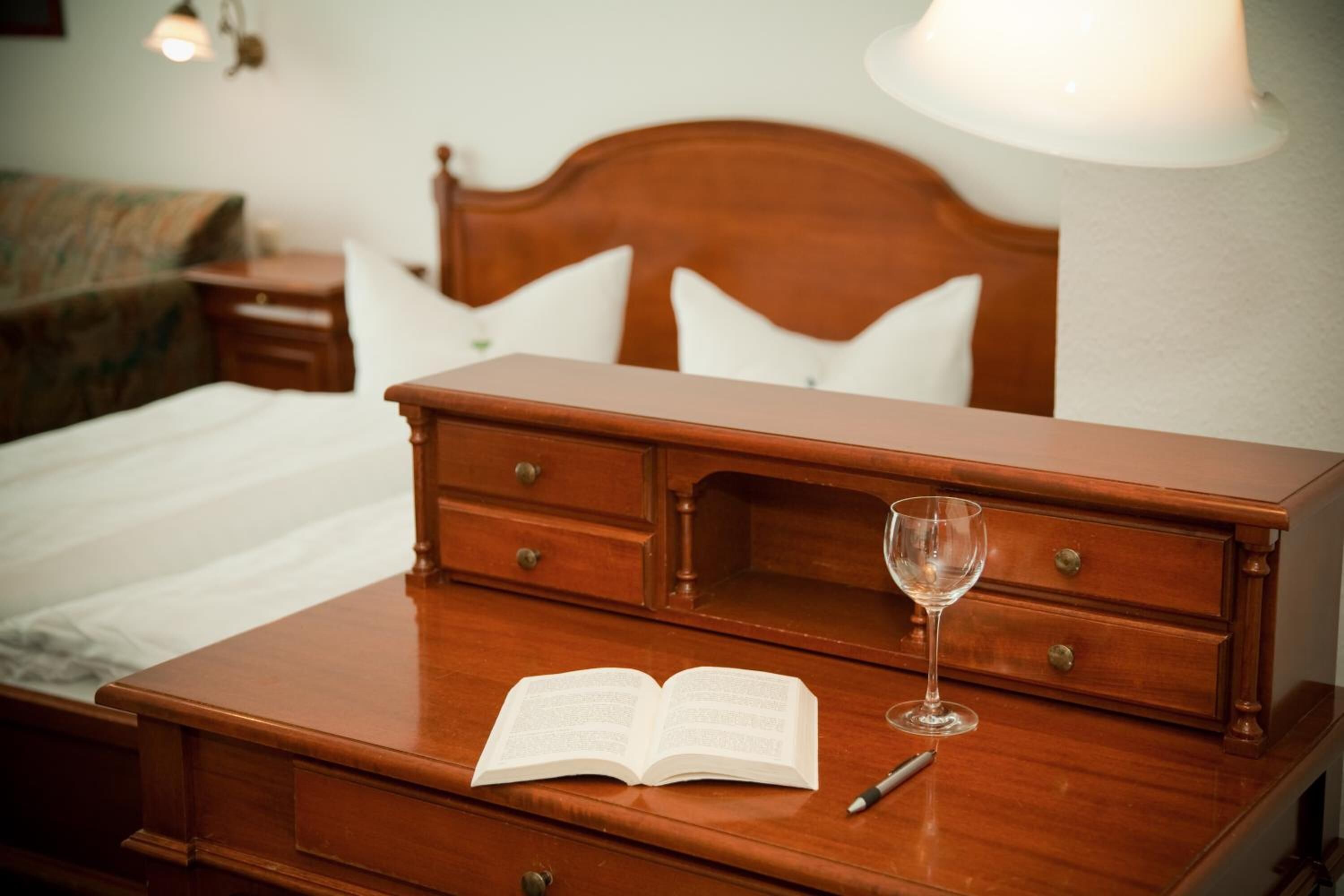 Ein Nachttisch aus Holz mit Buch und Weinglas, ein Bett mit weißer Bettwäsche und eine Wandlampe.