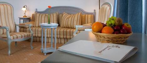 A room with a sofa, two chairs, a small table with a lamp, a basket of fruit, and a book on a table.