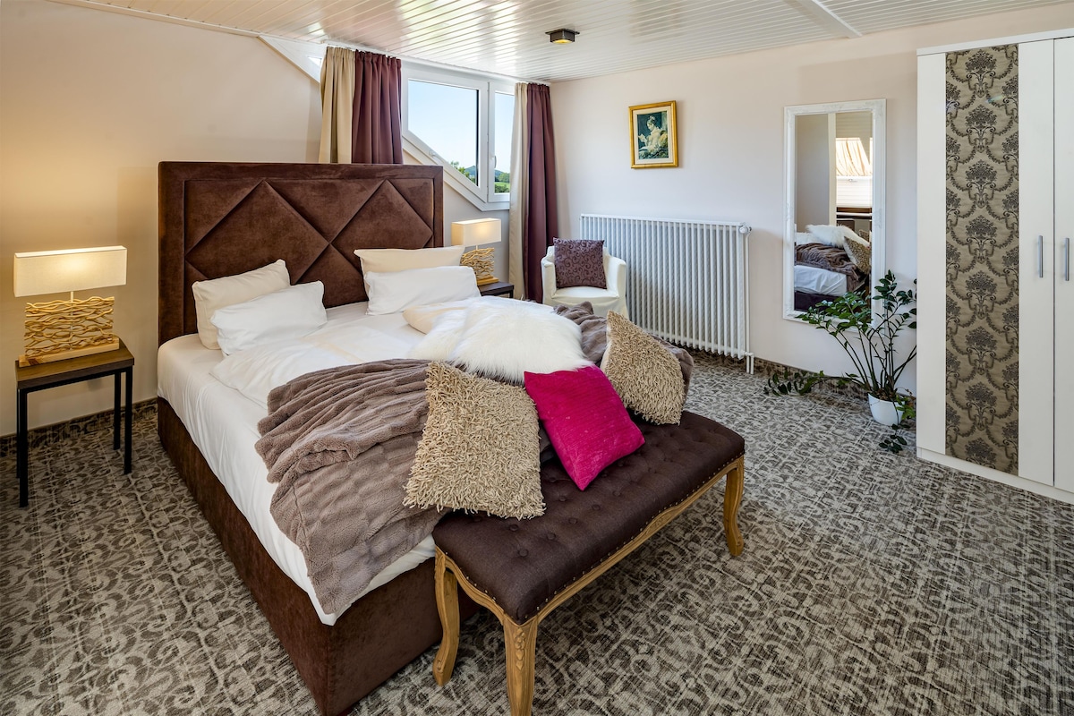 A well-appointed hotel room with a large bed, a sitting bench, a chair, a radiator, and a potted plant.