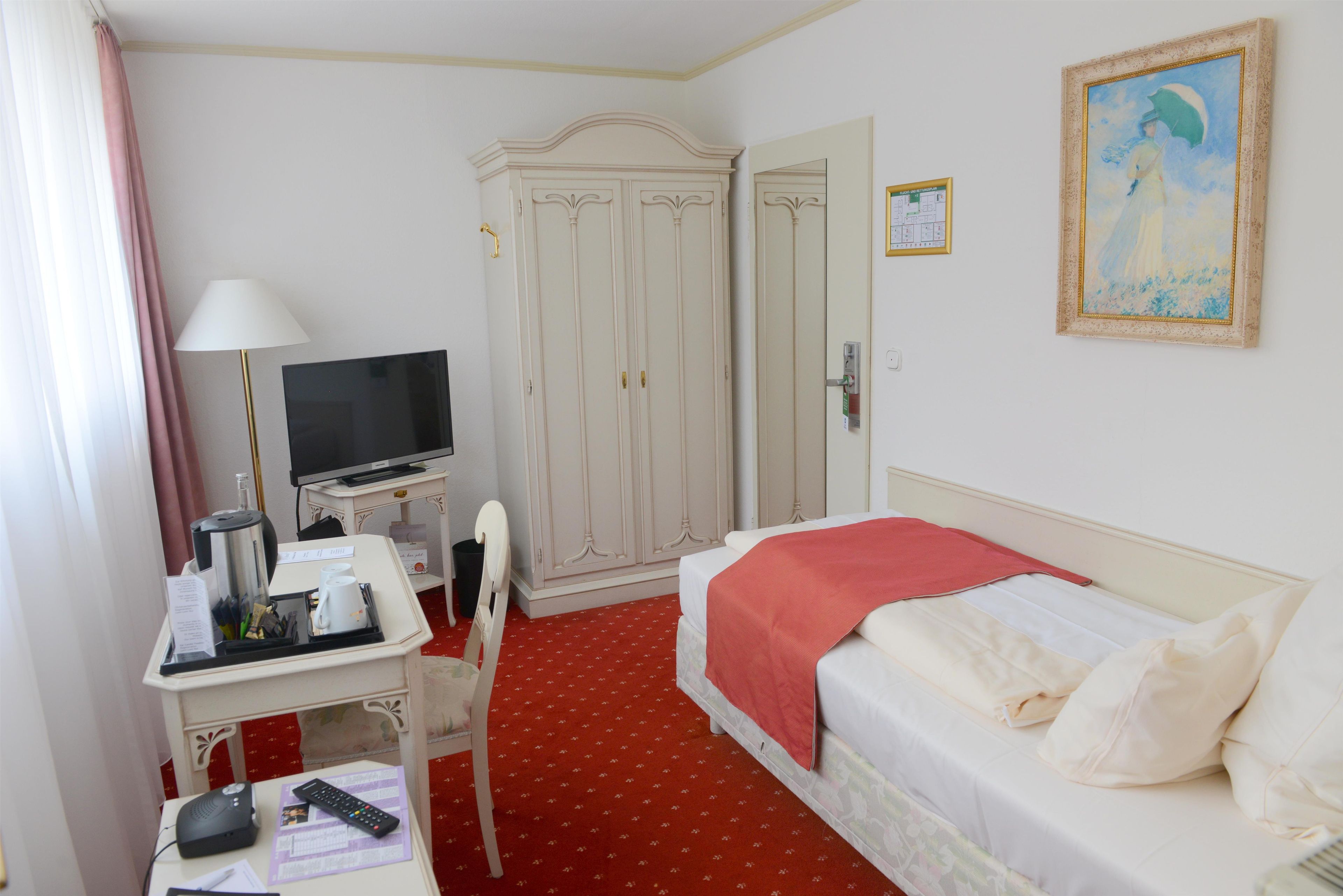 A hotel room with a bed, a desk with a TV, a wardrobe, and a painting on the wall.
