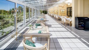 Een zonnige kamer met een glazen dak, grote ramen en een eethoek met tafels en stoelen.