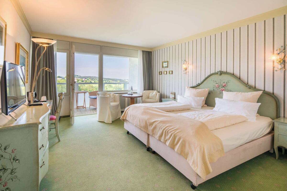 A hotel room with a large bed, a desk, a chair, and a balcony with a view.