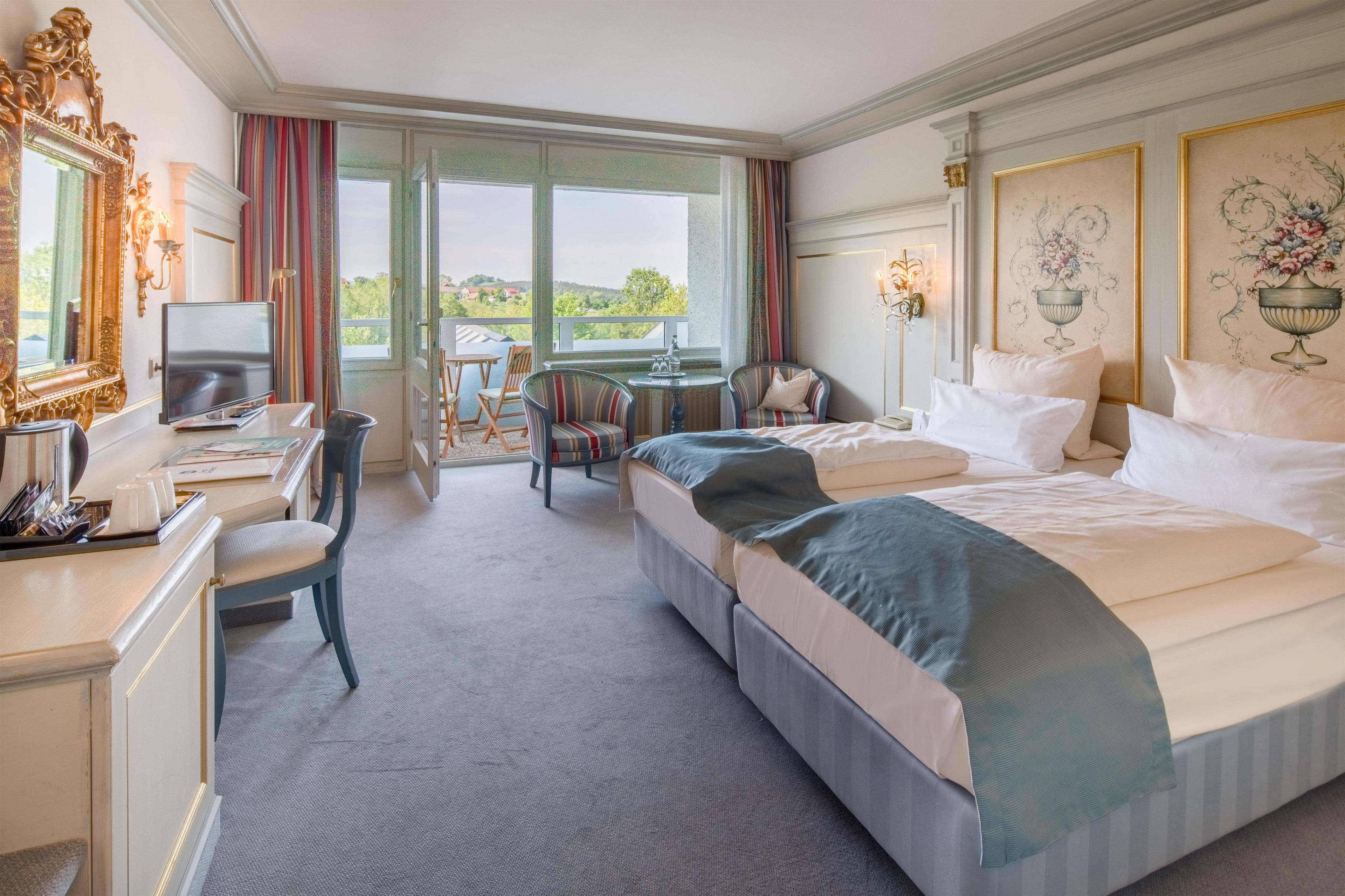 A hotel room with a bed, a desk, a chair, a small table, and a balcony with a view of the landscape.