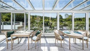 Een zonnige kamer met glazen dak, rieten meubels en uitzicht op groen.
