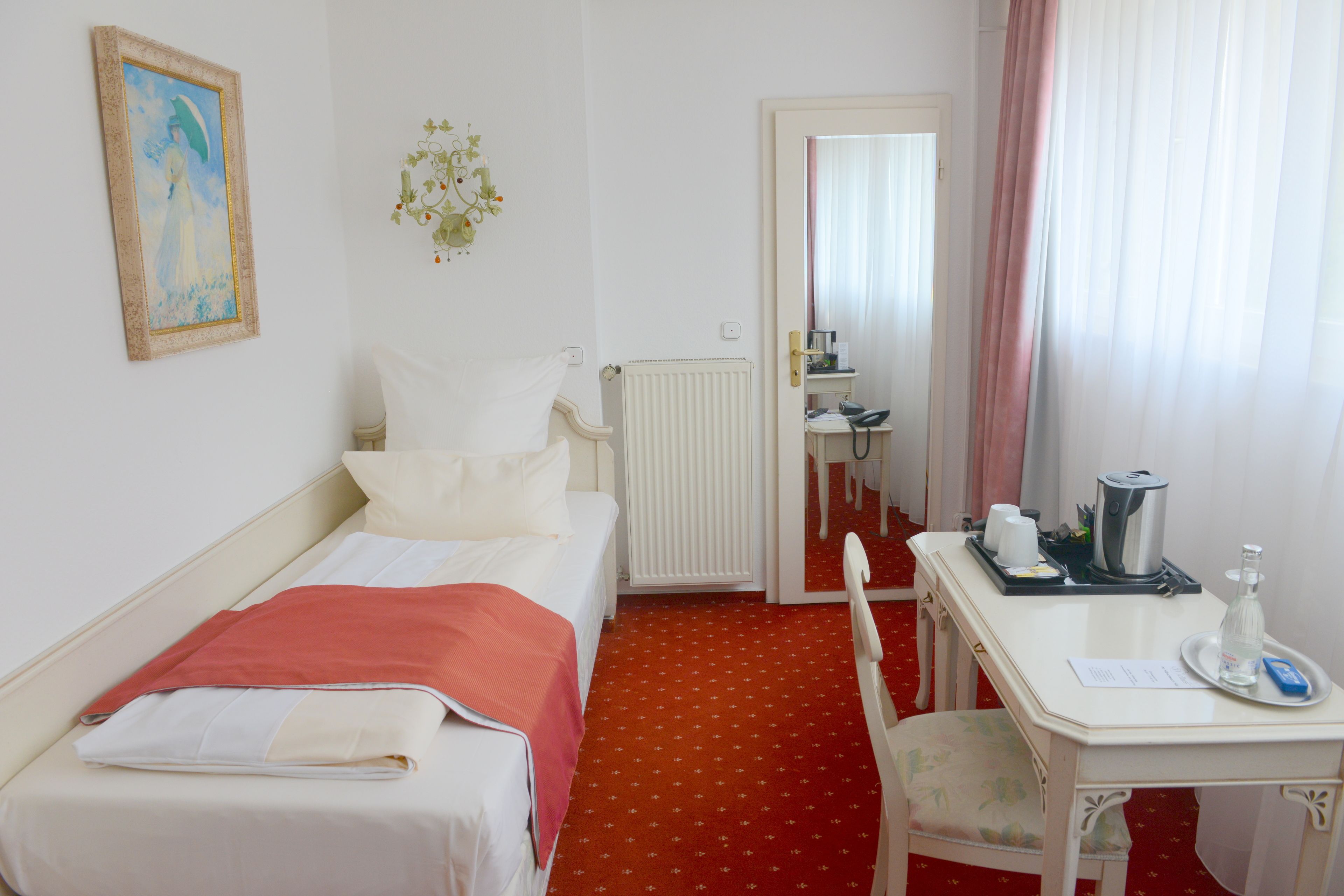 A hotel room with a bed, a desk with a chair, a radiator, a mirror, and a window with curtains.
