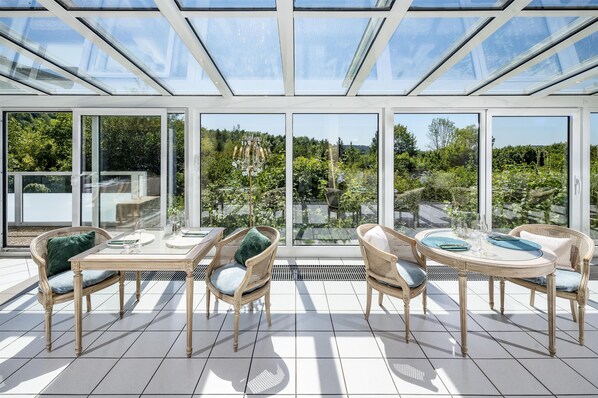 A sunlit patio with glass walls, wicker furniture, and a dining table set for four.