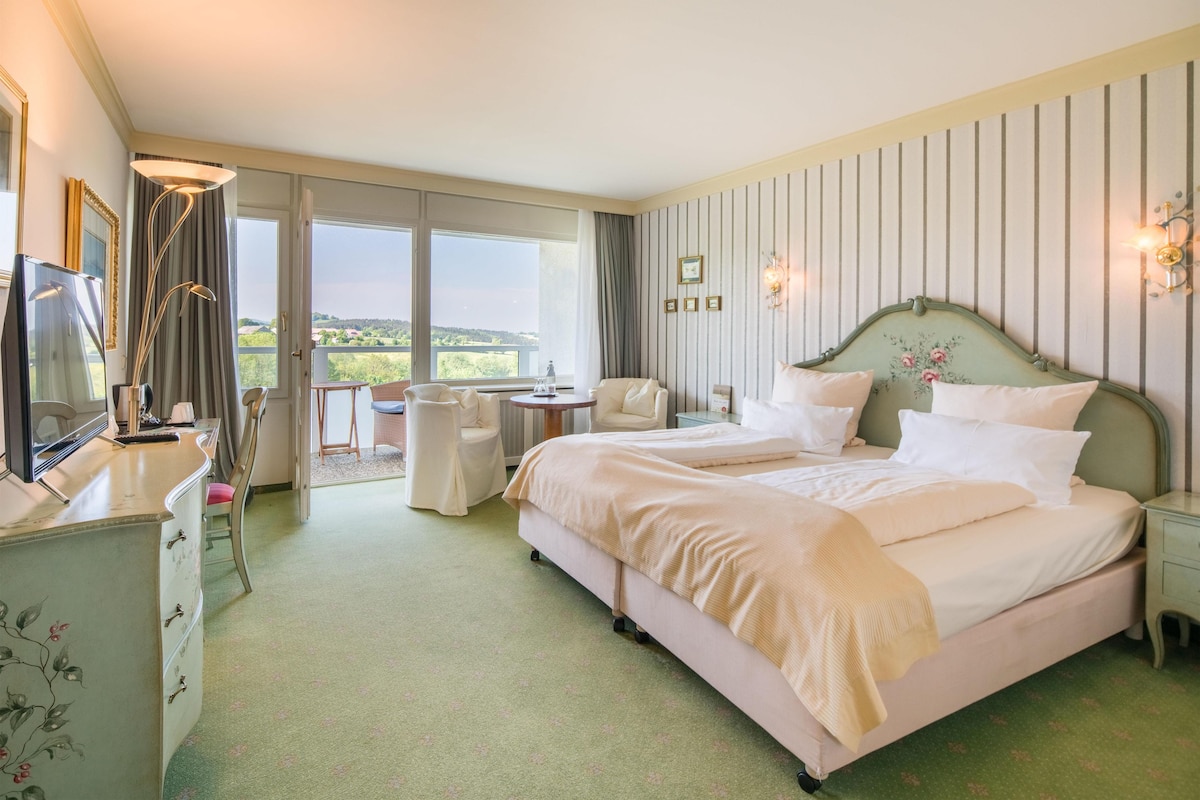 A hotel room with a large bed, a desk with a chair, a television, and a balcony with a view of the countryside.
