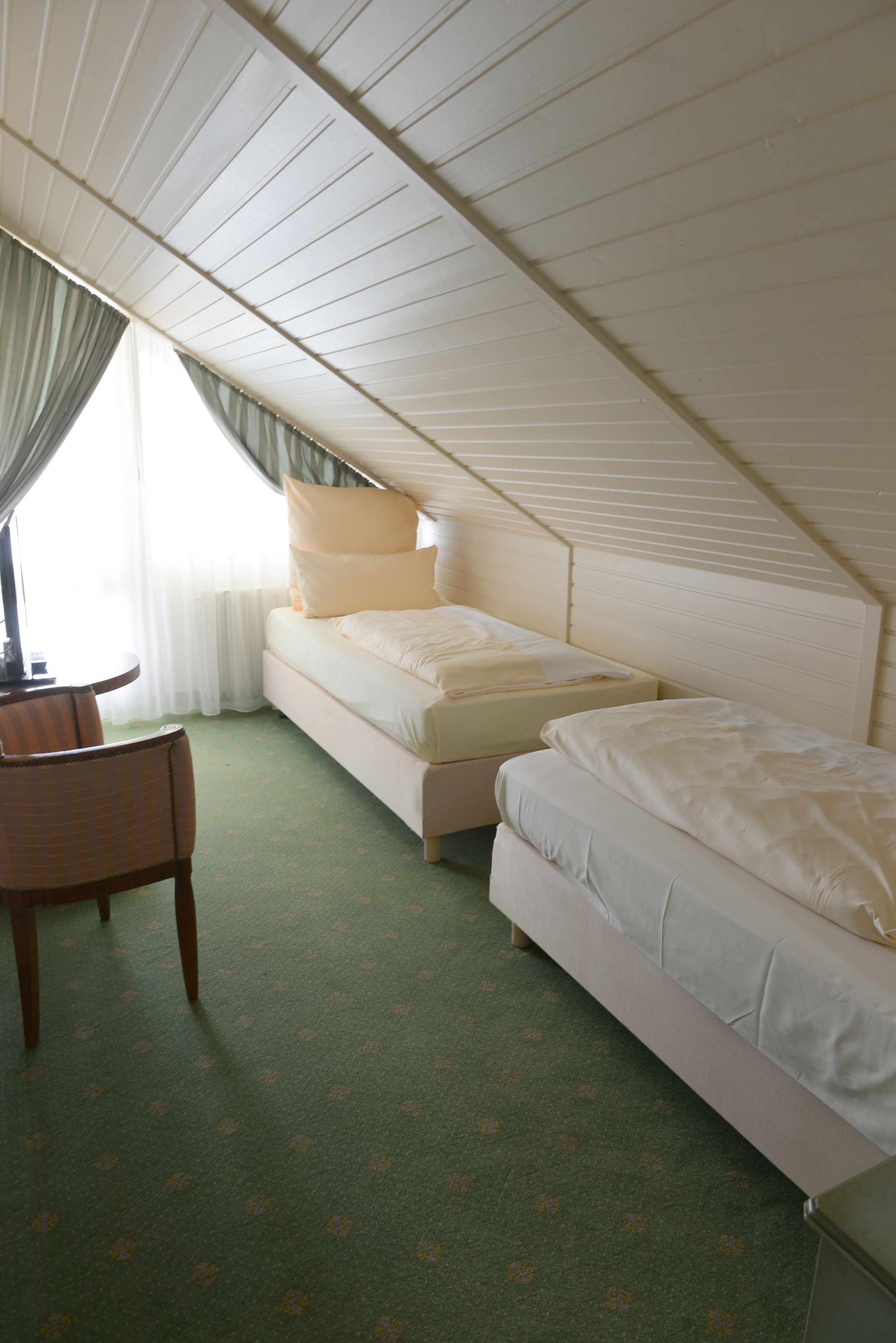 A small, single-room hotel room with a sloped ceiling, two beds, a chair, and a window with curtains.