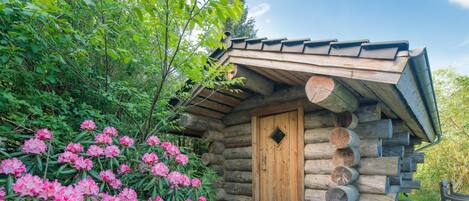 A rustic log cabin with a wooden door, surrounded by lush greenery and flowering bushes.