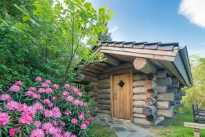 A rustic log cabin with a wooden door, surrounded by lush greenery and flowering bushes.