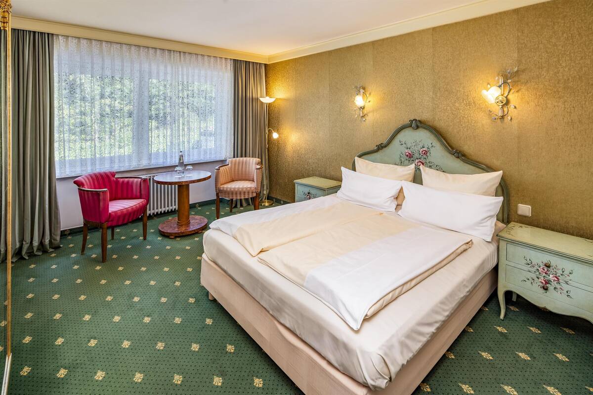 A hotel room with a large bed, a small round table, a red chair, and a nightstand with a floral design.