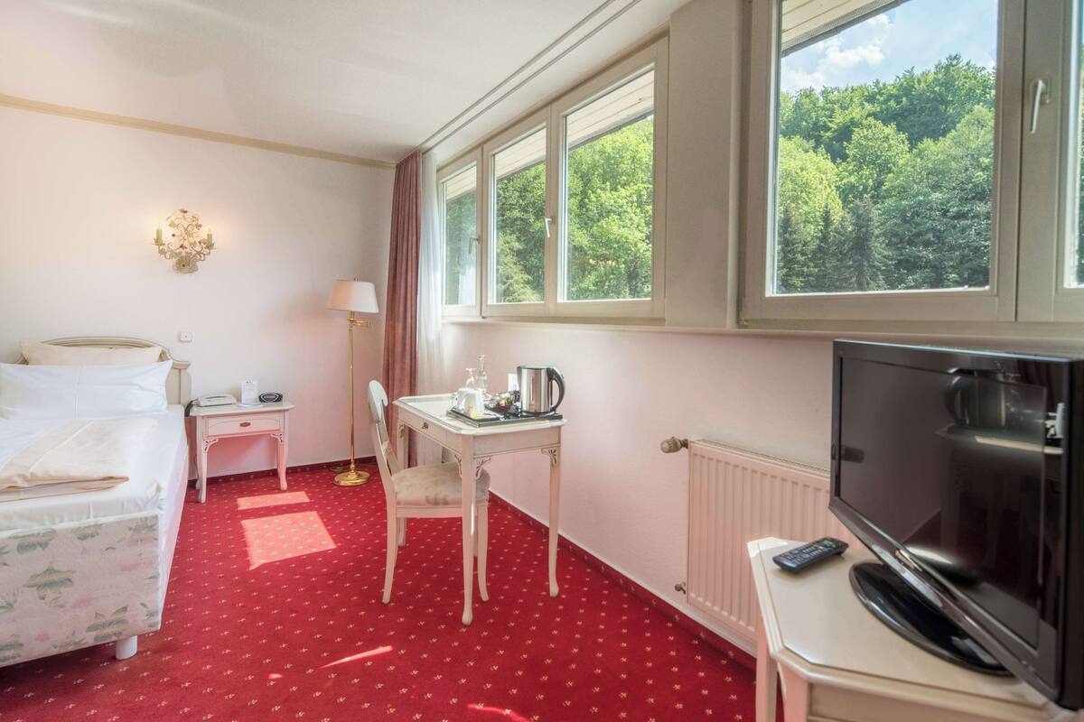 A hotel room with a bed, a desk with a chair, a television, and a window with a view of trees.