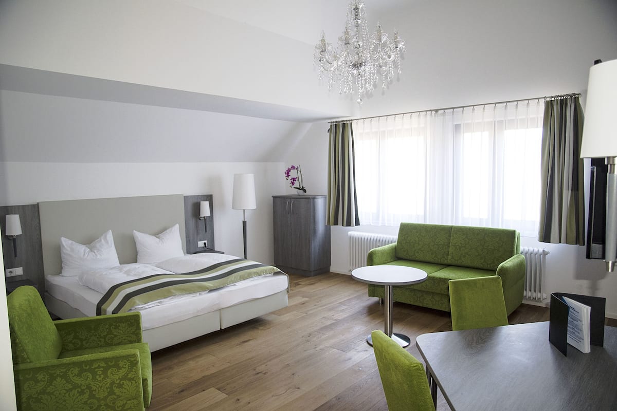 A hotel room with a bed, a green sofa, a small round table, a chandelier, and a radiator.