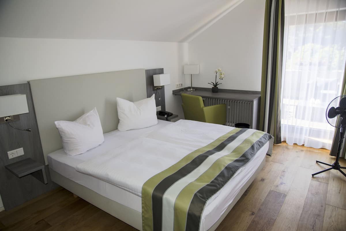 A hotel room with a bed, bedside table, lamp, chair, desk, and a window with curtains.