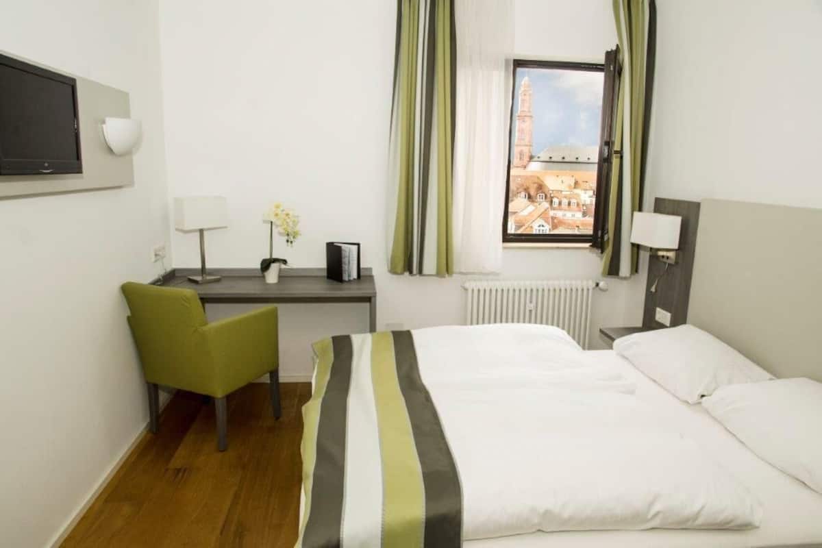 A hotel room with a bed, a desk with a chair, a TV, a window with curtains, and a radiator.