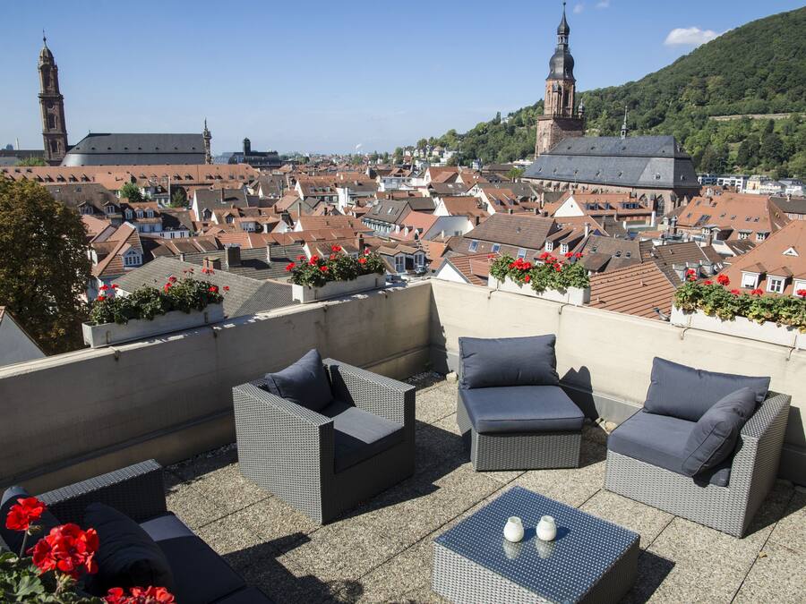 Una terraza en la azotea con mobiliario de mimbre y plantas en macetas, con vistas a un pueblo histórico con edificios destacados y una iglesia.