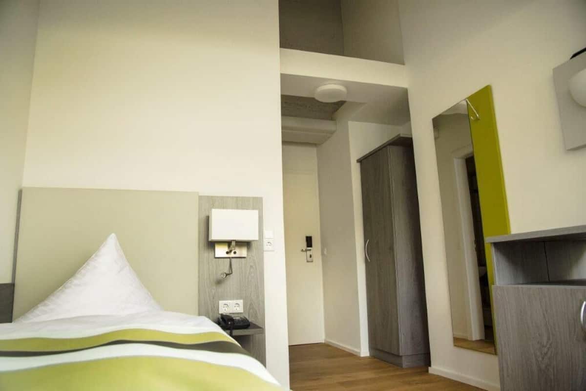 A hotel room with a bed, a bedside table with a phone, a wall-mounted light, and a wardrobe.