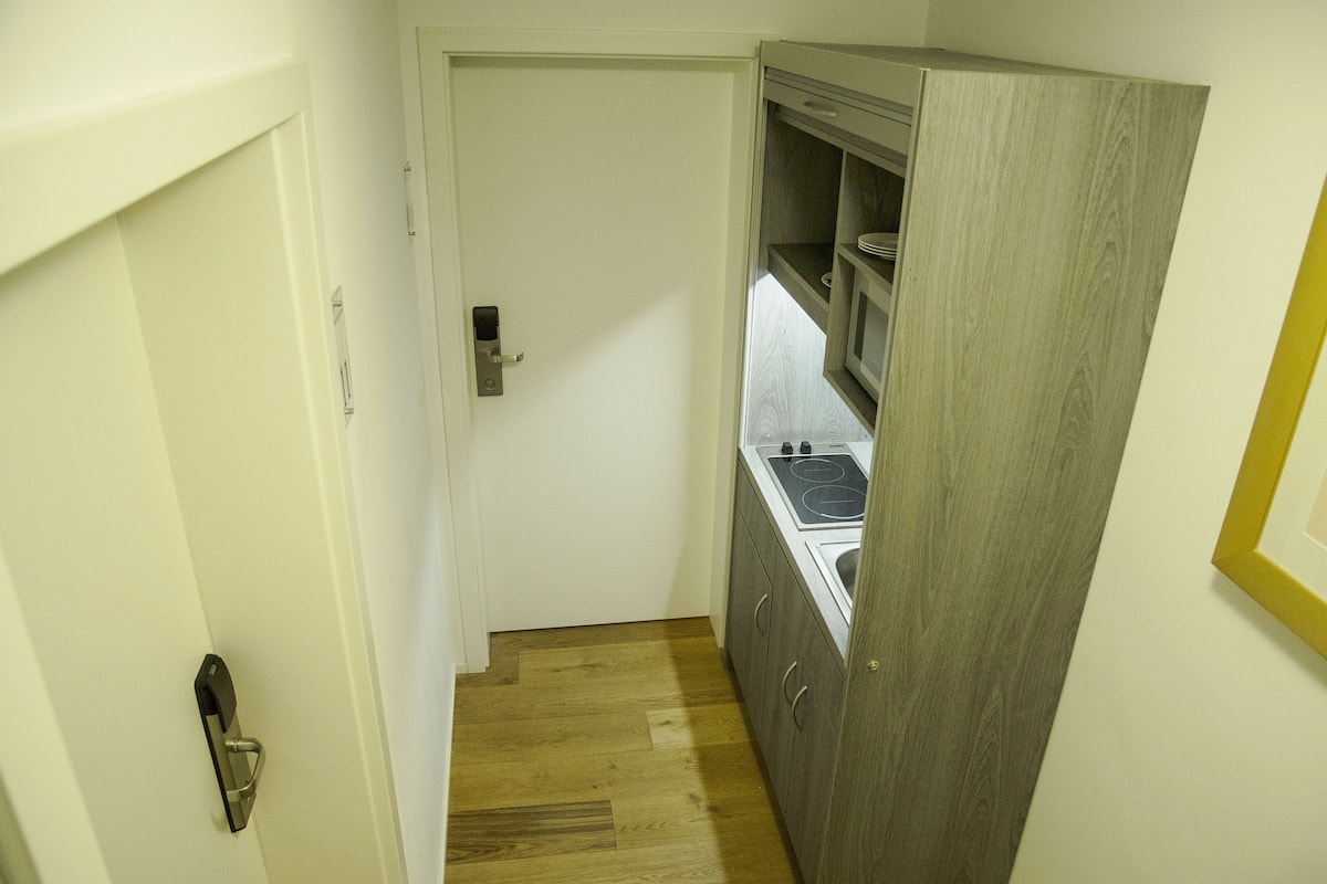 A narrow hallway with a white door, a wooden floor, and a kitchen area with a stove and cabinets.