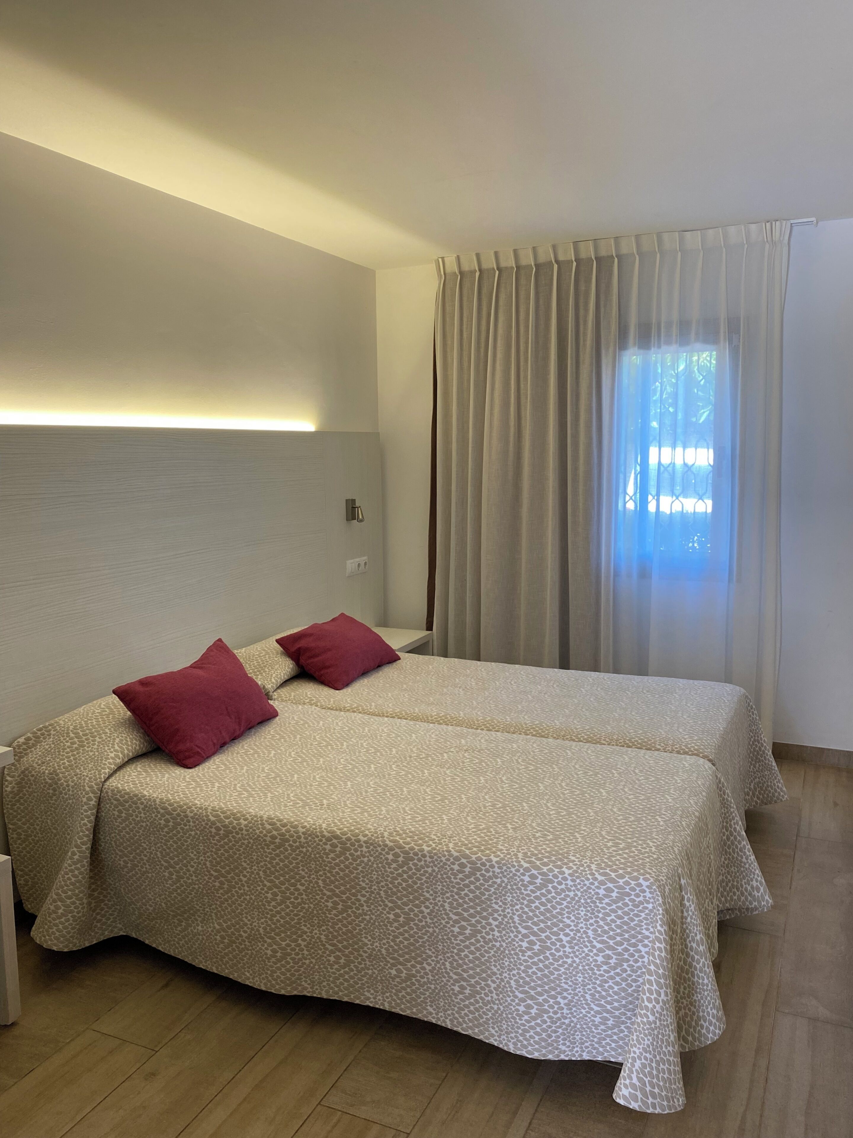 A hotel room with a bed, two pillows, a nightstand, a wall-mounted light, and a window with curtains.