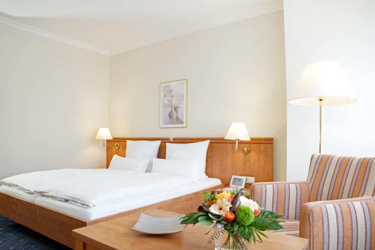 Habitación de hotel con una cama bien hecha, dos lámparas de noche, un cabecero de madera, una mesita con un jarrón de flores y un sillón a rayas.