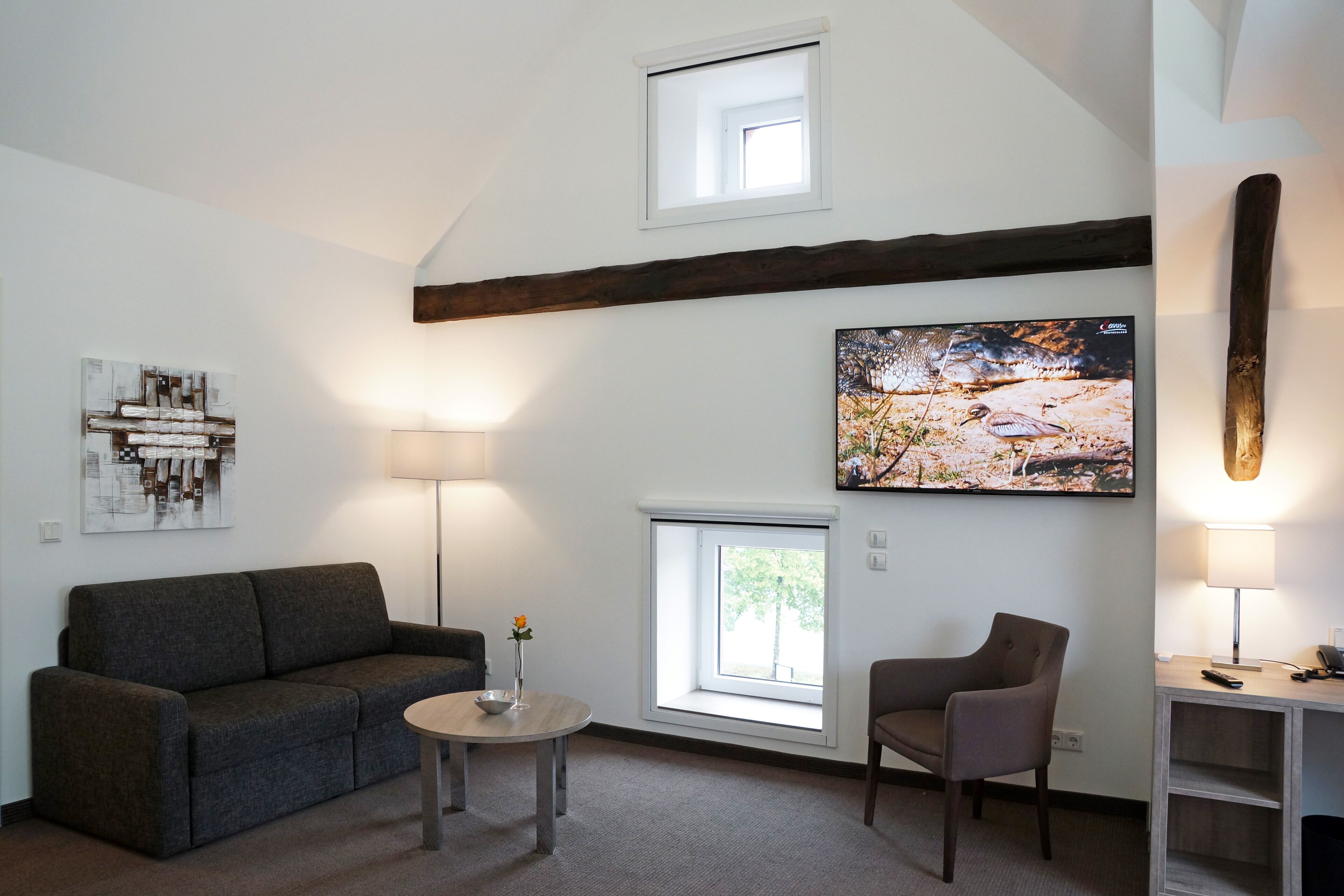 A modern living room with a gray sofa, a round table with a vase, a chair, and two windows.