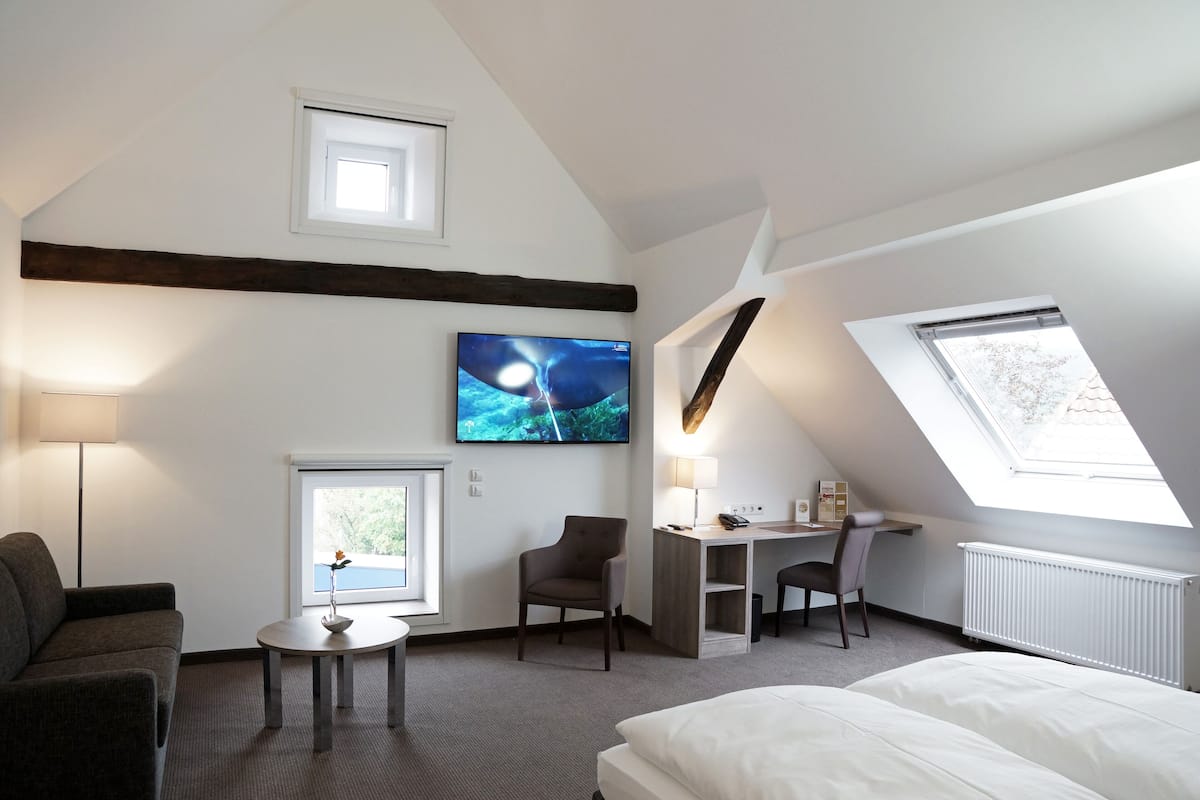 Una habitación de hotel moderna con techo inclinado, una cama grande, un escritorio con silla, una pequeña mesa redonda con un jarrón y un televisor montado en la pared.