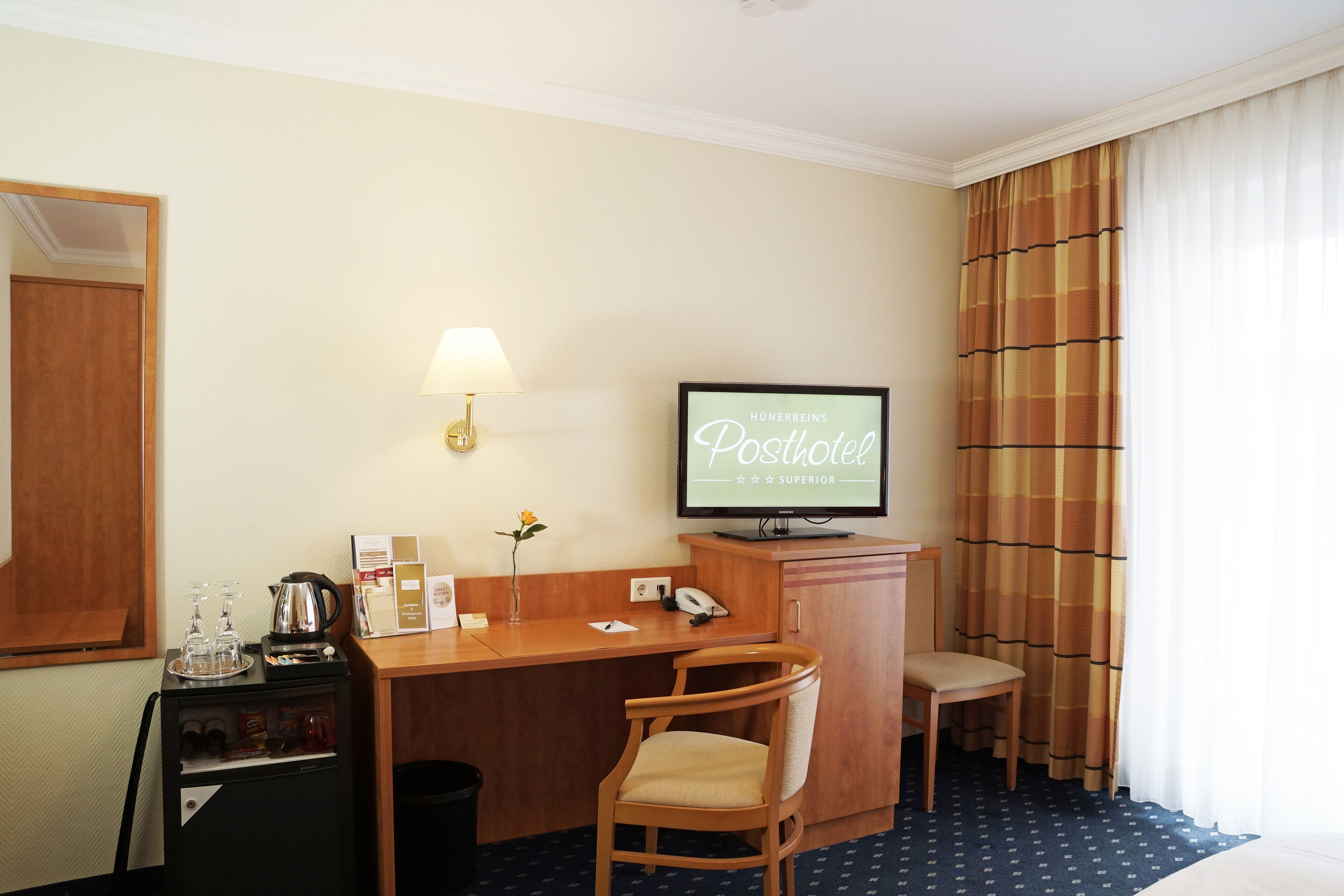 Una habitación de hotel con un escritorio, un televisor mostrando 'Hunerbeins Postoffice', una silla, un mueble bajo con una tetera y un espejo.