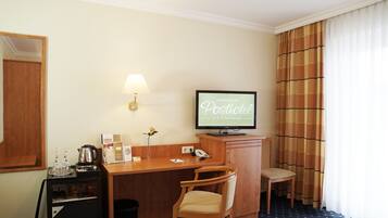 A hotel room with a desk, a television displaying 'Hunerbeins Postoffice', a chair, a small cabinet with a kettle, and a mirror.