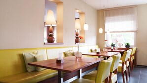 A restaurant with wooden tables and yellow chairs, a yellow sofa, and a large window with blinds. - Hünerbein's Posthotel (Bissendorf)