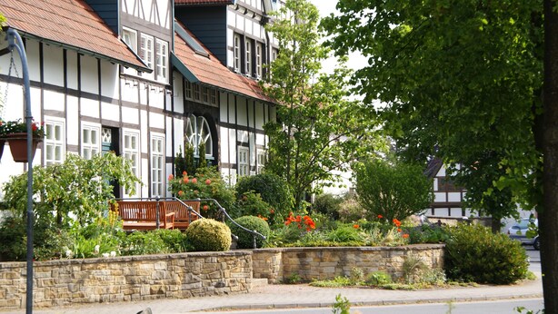 A traditional half-timbered house with a stone wall garden, a bench, and a car parked on the street. - Hünerbein's Posthotel (Bissendorf)