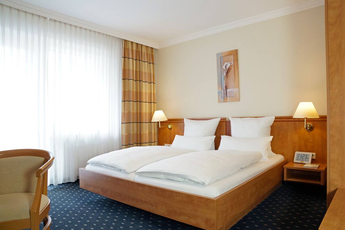 Habitación de hotel con una cama doble, dos mesitas de noche, una silla, un reloj y un cuadro en la pared.