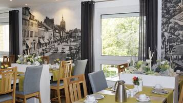 A dining area with wooden chairs and tables, a large mural on the wall, and a view of greenery outside.