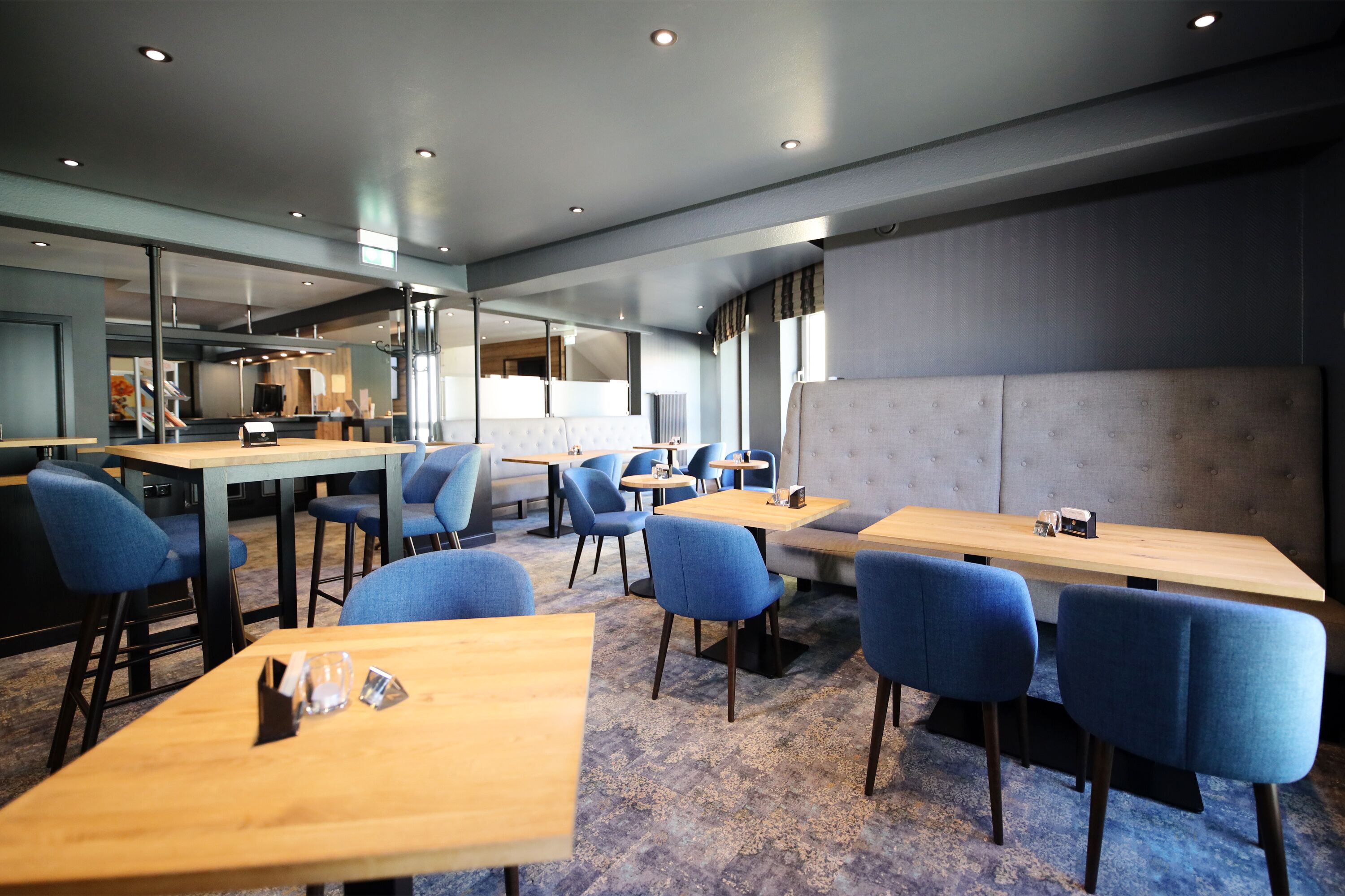 En modern restaurang med träbord och blå stolar, en grå, vadderad bänk och ett stort fönster som släpper in naturligt ljus.