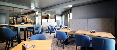 Un restaurant moderne avec des tables en bois et des chaises bleues, un banc rembourré de couleur grise et une grande fenêtre laissant entrer la lumière naturelle.